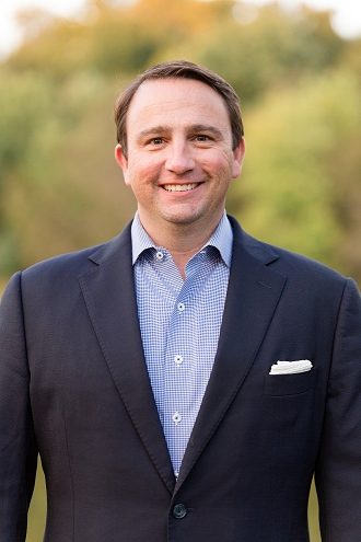 Portrait of a smiling man dressed in a business suit, owner of C&S Cabinets, standing outdoors with a blurred natural backdrop.